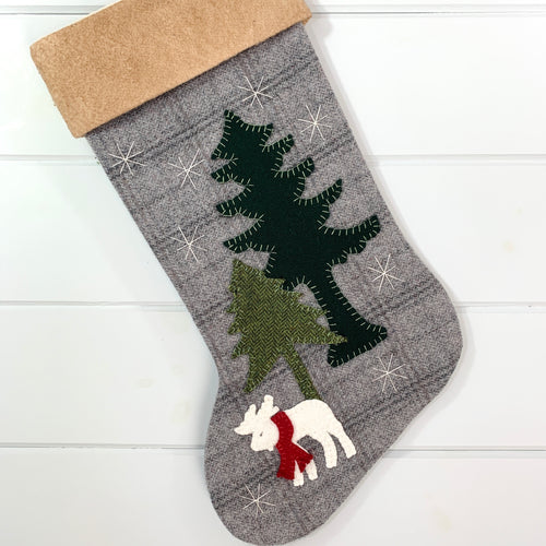 winter white moose Christmas stocking, moose is wearing a red wool fabric scarf around neck, moose is white wool fabric with gray  stitched eye, two pine trees, one is tall dark green wool fabric, one is short green plaid wool fabric, white stitched snowflakes, stocking is made of gray plaid wool fabric, cuff is tea dyed cotton batting, woodland Christmas stocking, rustic Christmas stocking