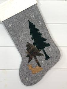 baby deer Christmas stocking, fawn Christmas stocking, stocking is gray herringbone wool fabric, fawn, pine trees are hand stitched to stocking, fawn is light brown wool fabric with white stitched spots on its back, tall pine tree is dark green wool fabric, short pine tree is green plaid wool fabric, white snowflakes are hand stitched onto stocking, cuff is white natural cotton batting, woodland Christmas stocking, baby deer rustic Christmas stocking