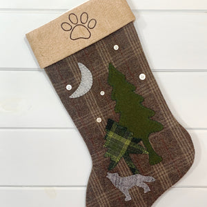 paw print Christmas stocking, brown plaid wool fabric, cuff made from tea dyed cotton batting, cuff has a black  stitched paw print of a dog or animal, stocking has gray wool fabric wolf howling at a light gray wool fabric moon, two pine trees, one green wool fabric, one green plaid wool fabric,  five white vintage buttons used for snowflakes, large stocking, woodland forest Christmas stocking